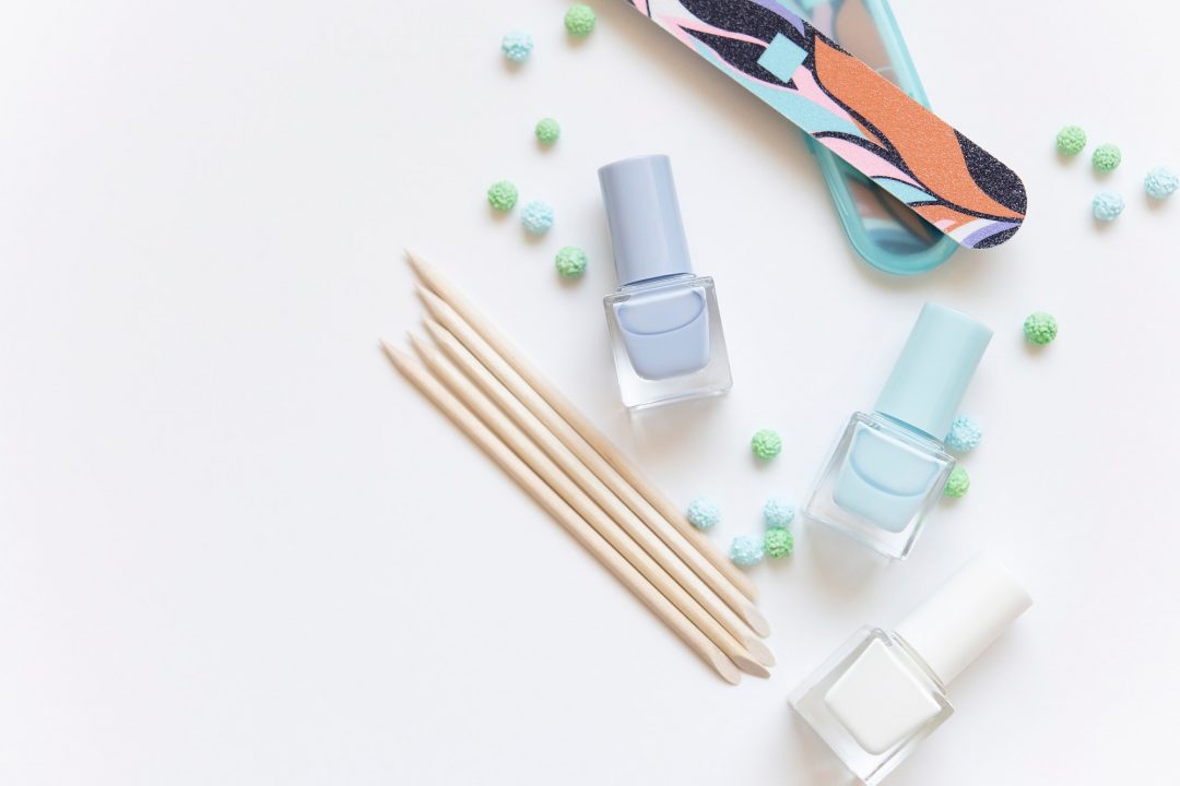 Un conjunto de herramientas para manicura sobre un fondo blanco, incluyendo tres frascos de esmalte en tonos pastel, una lima decorativa y palitos de madera, junto a pequeñas decoraciones esféricas verdes y azules.
