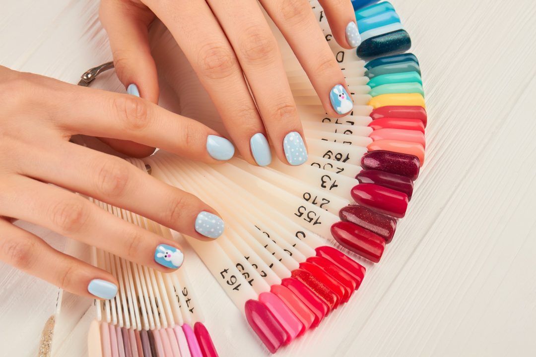 Una mano con uñas pintadas de azul claro con detalles decorativos, incluyendo puntos blancos y un diseño de muñeco de nieve, sostenida sobre un abanico de muestras de uñas en tonos vibrantes y numeradas.